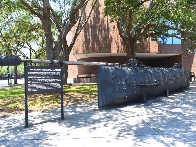 A replica of the H.L. Hunley Civil War submarine sits outside the brick exterior of the Charleston Museum, shaded by large trees