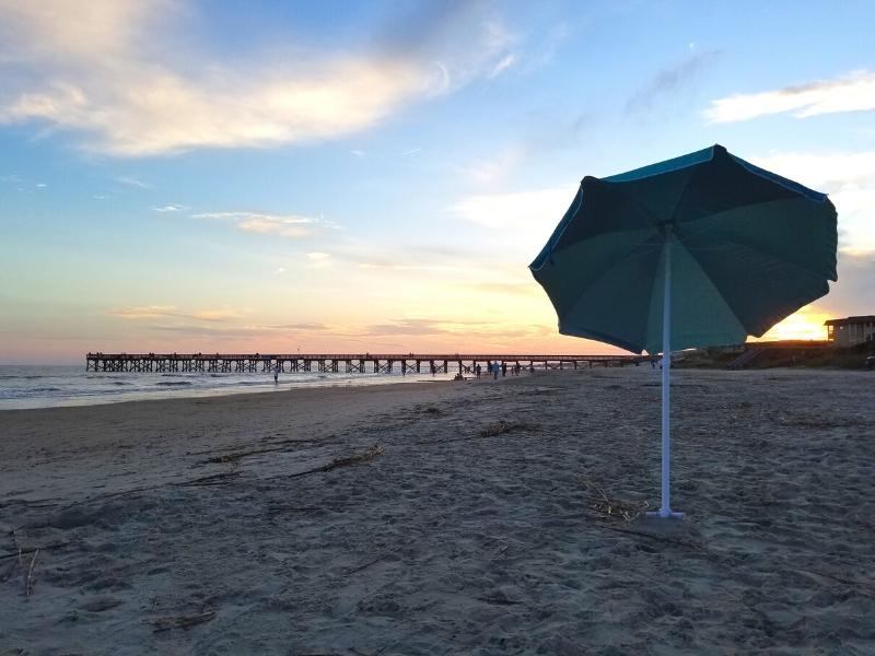 A lone beach umbrella stands with Isle of Palms pier in the background as the sun sets