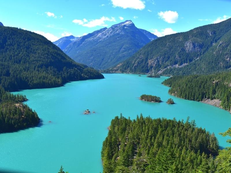 Chalky teal blue waters of Diablo Lake glow on a clear day, surrounded by green forested mountainsides and a few tiny islands in the lake