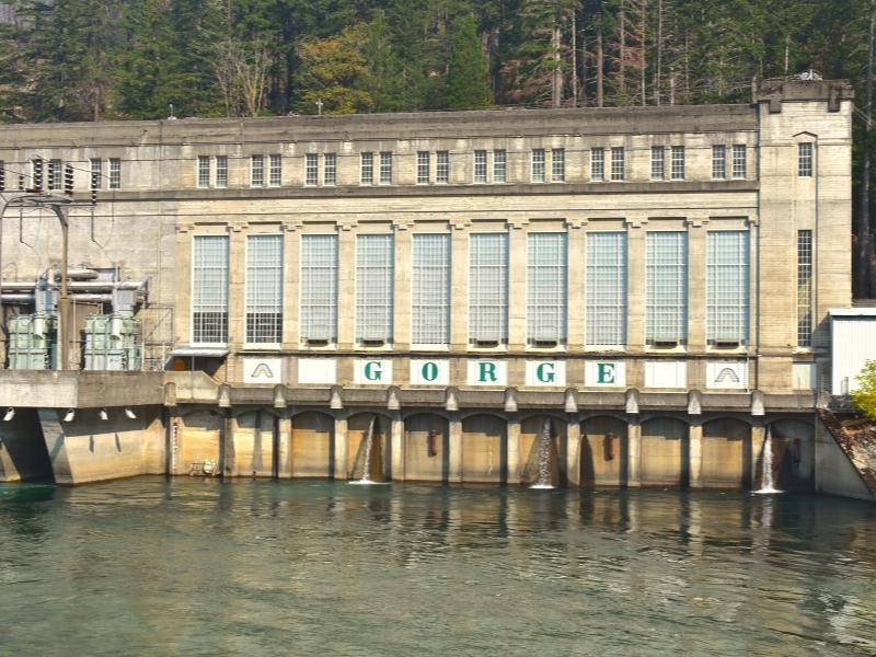 The tan stone facade of the Gorge Powerhouse with rows of tall windows and the Skagit River in the foreground