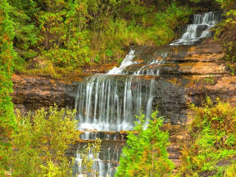 Alger Falls drops several tiers down a rocky cliff, surrounded by green summer foliage