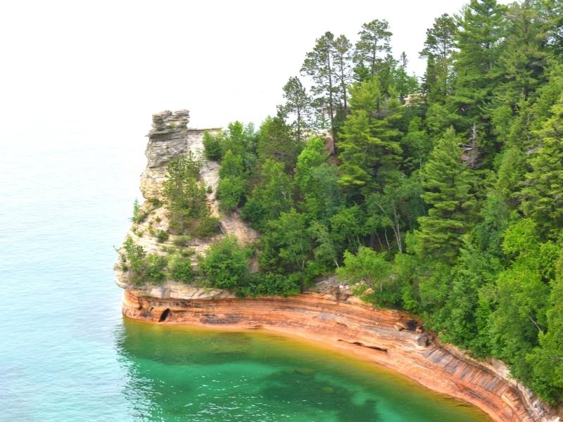 The Miners Castle cliff rock formation abuts green trees and the blue-green Lake Superior with vibrant orange cliffs on a gray day