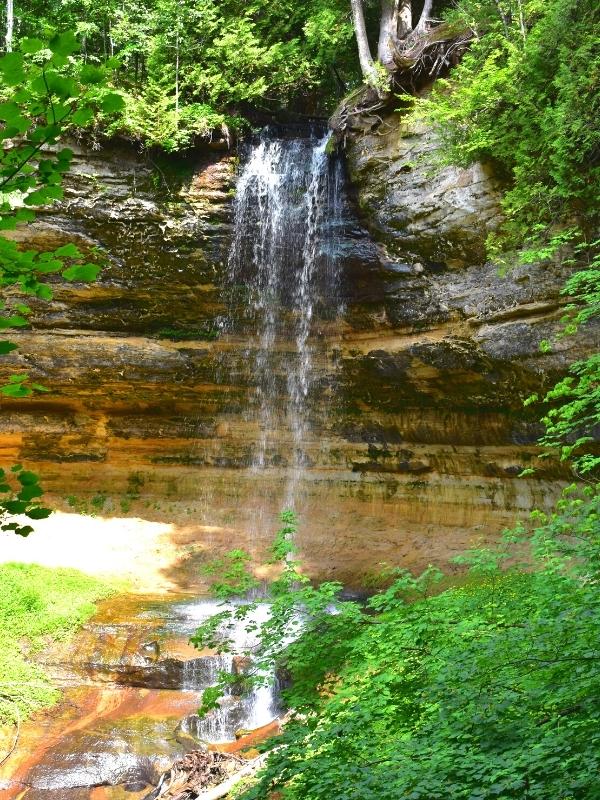 Pictured Rocks' Munising Falls steadily erodes the gorge it drops into, surrounded by green trees in summer