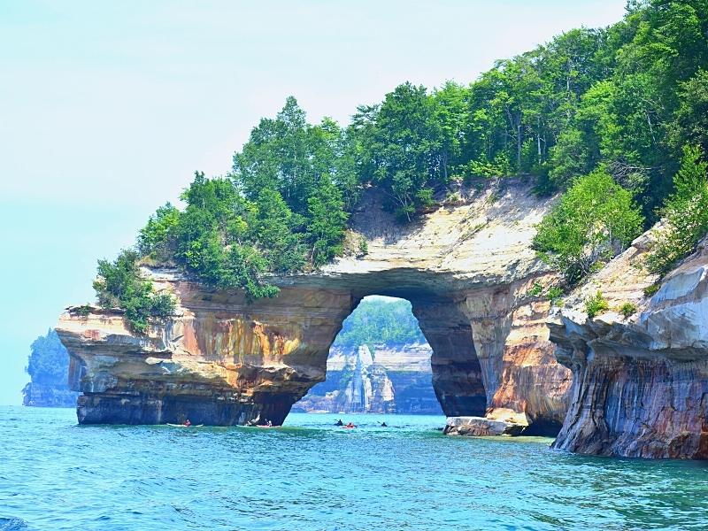 A large archway is eroded into the cliffs of Pictured Rocks, with green trees above it and the blue-green waters of Lake Superior below it.