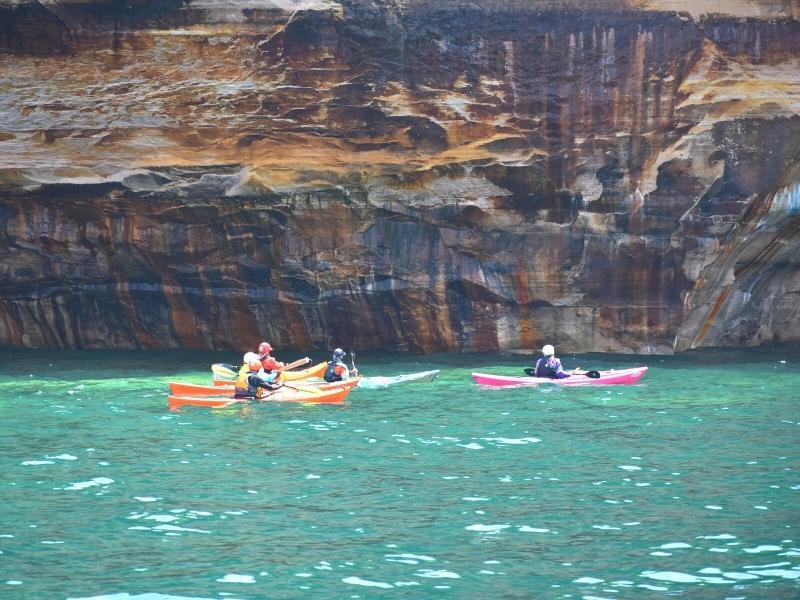 Kayakers in colorful boats float on the aqua green waters of Lake Superior next to the tan and black cliffs of Pictured Rocks