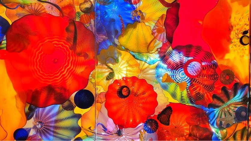 Reds, yellows, and blues in several circular patterns, all piled on top of each other and displayed in a ceiling through clear glass at the Chihuly museum in Seattle