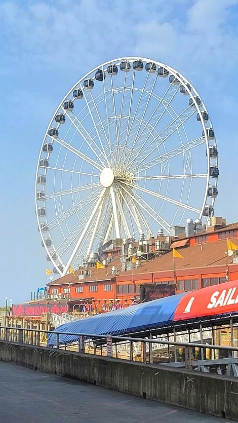 A large white Ferris wheel, the Seattle Great Wheel, rises above the roofs of the wharf on a semi-cloudy day