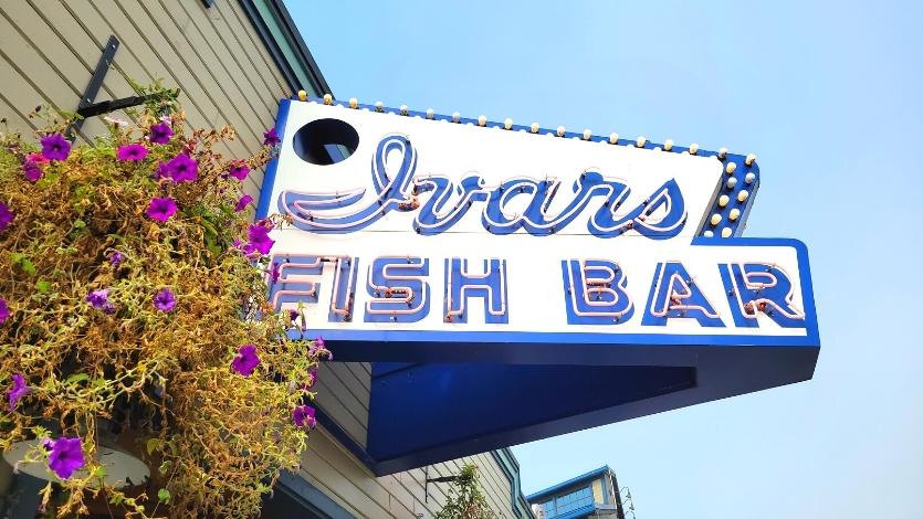 Neon blue and white sign for Ivar's Fish Bar, attached to the siding with a hanging flower pot