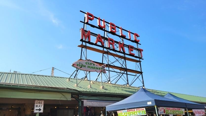 A neon sign for Public Market and a fish neon sign for City Fish Market sit atop the green metal roof of Pike Place Market in downtown Seattle