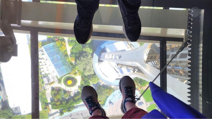 Feet in sneakers stand on top of the Space Needle's glass floor, with the Chihuly Gardens seen on the ground below.