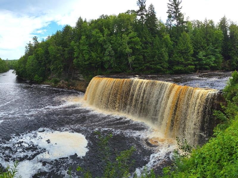 Surrounded by green forest, tan-colored Upper Tahquamenon Falls cascades over a cliff creating foam in the water