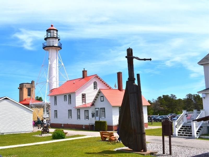 Several white buildings with a white lighthouse in the background and a wooden ship rudder at the Great Lakes Shipwreck Museum complex at Whitefish Point