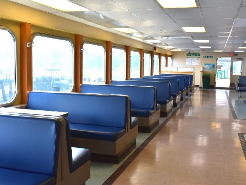 The interior cabin of a Whidbey Island ferry features large windows with blue upholstered benches with tables.