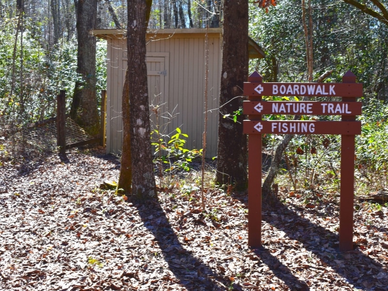 A brown trail sign indicates the boardwalk, nature trail, and fishing are all to the left, with a small tan maintenance shed in the background.