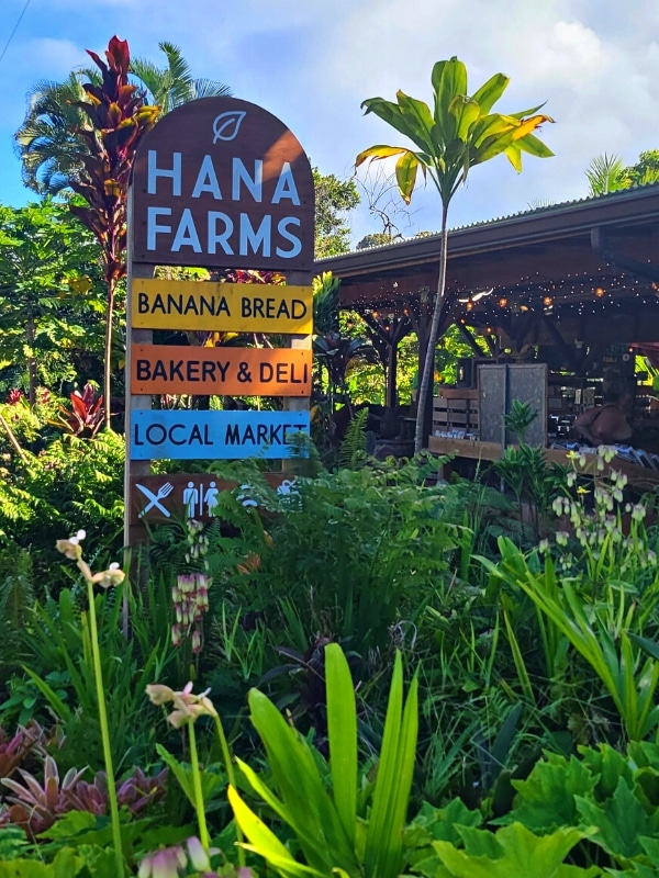 A sign for Hana Farms' banana bread, bakery & deli, and local market sits in colorful vegetation by the store.