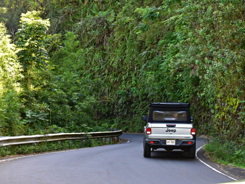 A gray Jeep navigates around a tight curve on the Road to Hana, completely surrounded by green cliffs.
