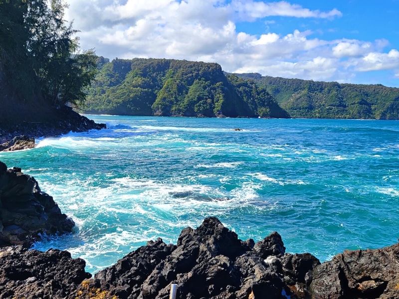 The Road to Hana's green forested cliffs surround a bay of blue and aqua as they crash on the black rocky coast.