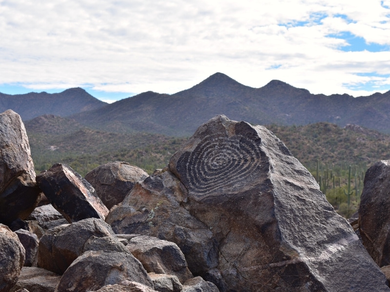 A circular swirl petroglyph adorns a large rock atop Signal Hill in Saguaro West with the Tucson mountains in the background.