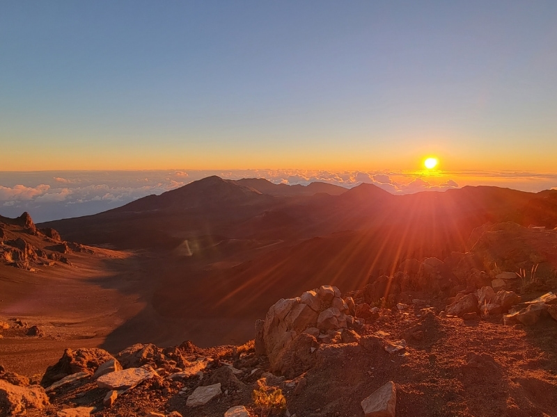 A bright orange sun sends out its rays over the Haleakala crater as the sun rises above the cloud bank