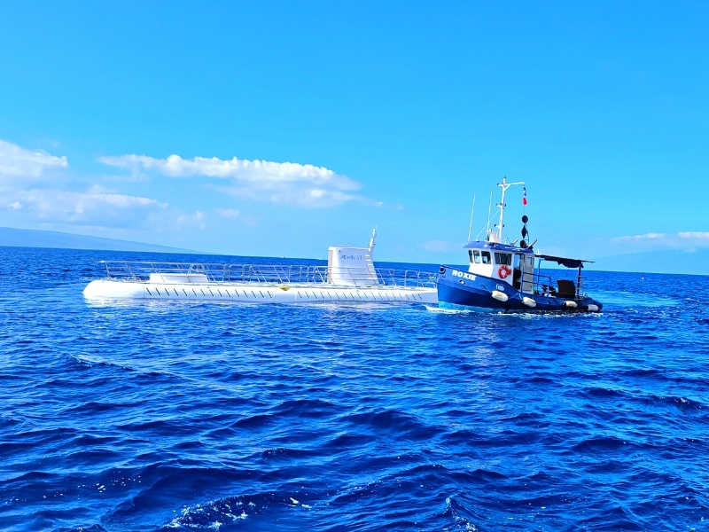 A small blue tugboat comes around the white hull of the Atlantis submarine in Maui