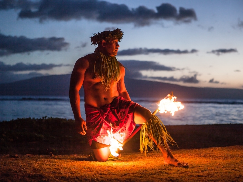 A man in native Hawaiian dress with a fire stick on the beach just after sunset
