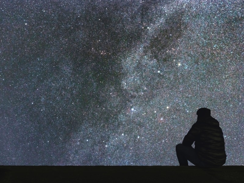 A man's shadow sitting on a curb with the starry night sky above him