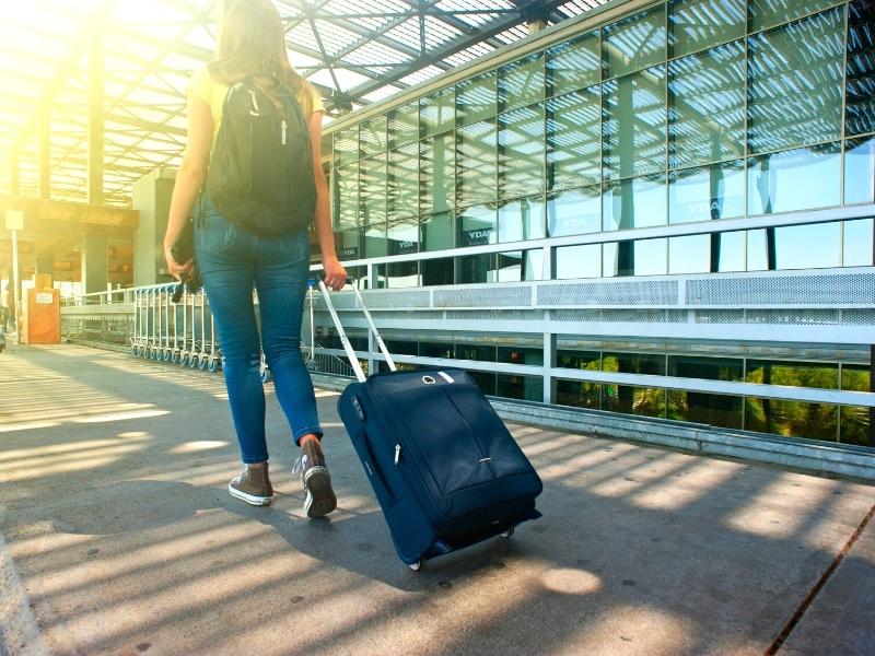 A woman with a backpack pulling a roller carry-on bag walks into the sun at an airport