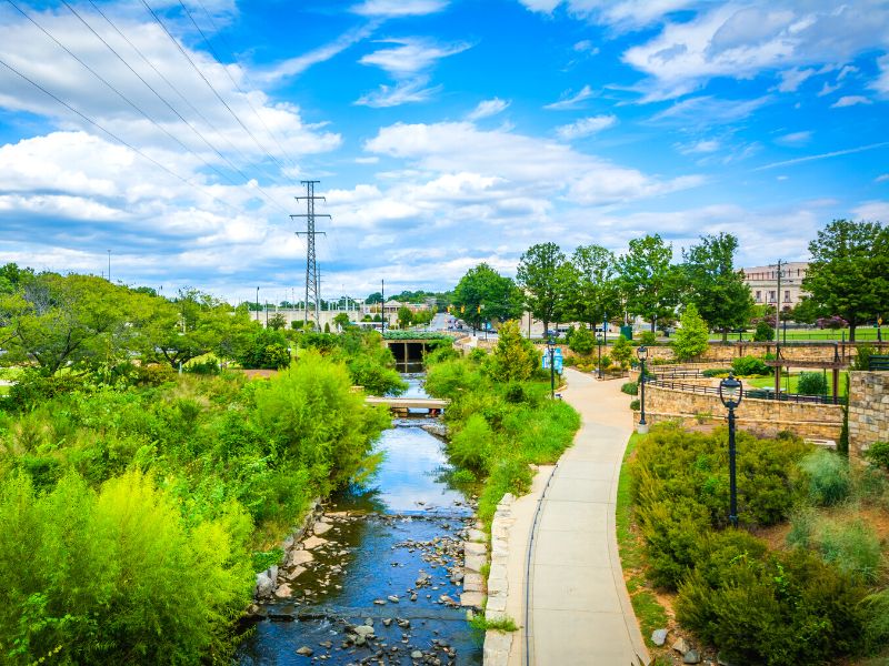 A paved path winds beside Little Sugar Creek under a blue sky with a landscaped park to the side.