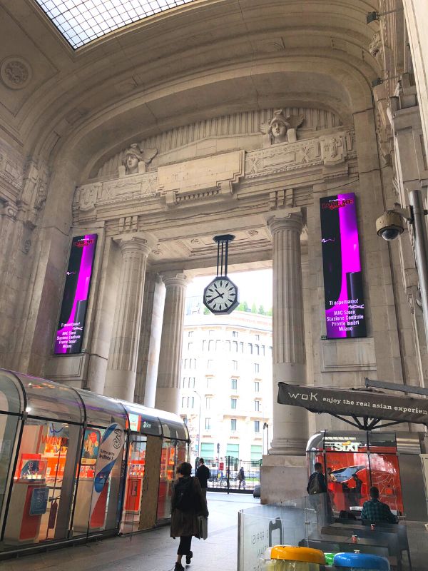 Inside the beautiful Milan Central Station, showing ornately carved moldings, columns, and an analog clock handing in the exit archway