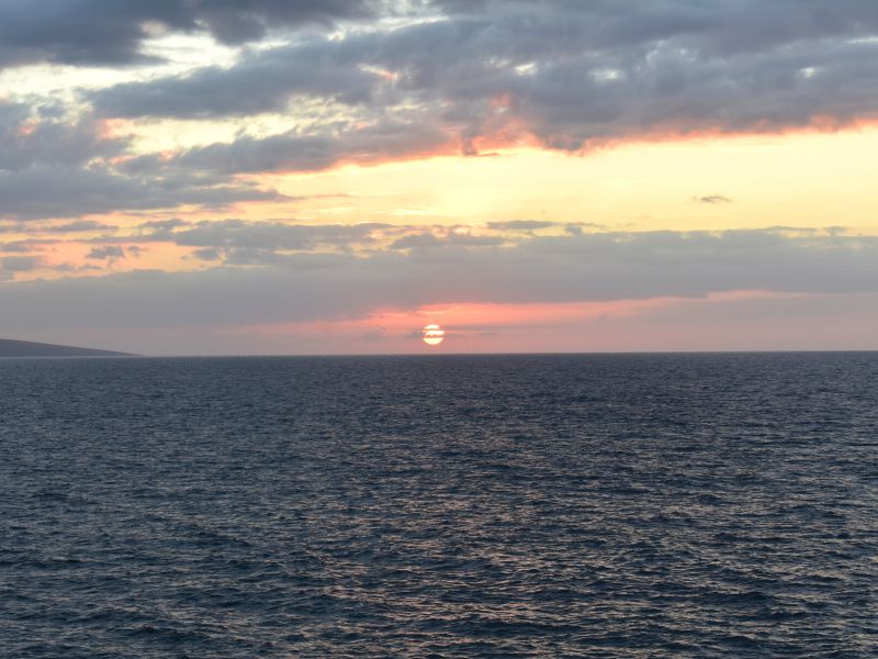 A fiery red orange sun sits just above the horizon with clouds and sky shades of pink, orange, and green at sunset on Maui