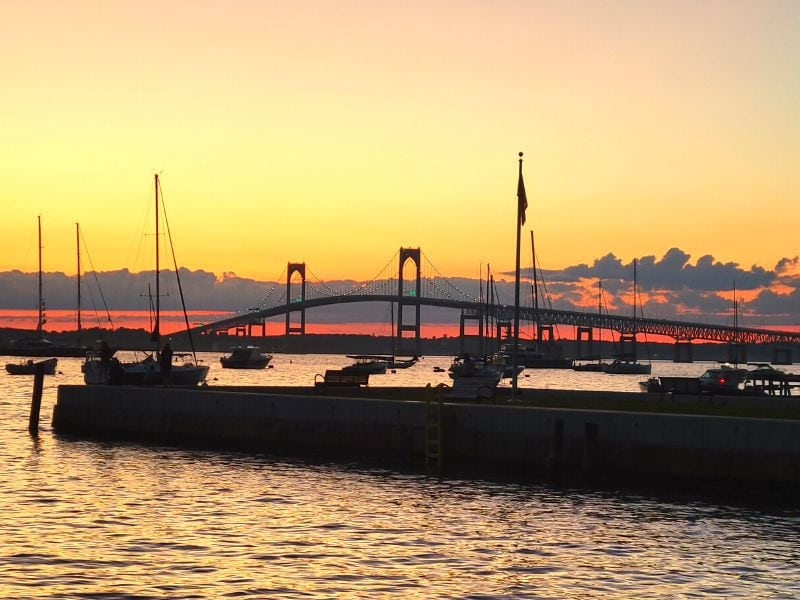 Shades of orange sunset color the sky behind the Newport bridge and harbor