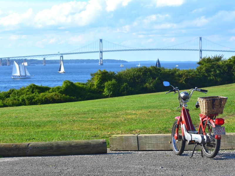 A moped parked in a lot overlooks Newport harbor and the bridge on a sunny day