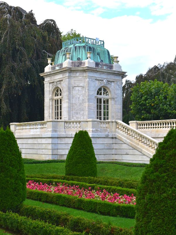 A garden folly with patina green roof, white stone, and surrounded by greenery and a hint of pink flowers at The Elms in Newport