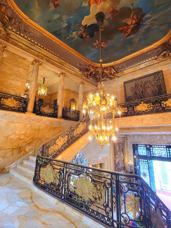 The front staircase in Newport, RI's Marble House, featuring floor to ceiling marble, gilded railings, muraled ceiling, and an enormous gold chandelier